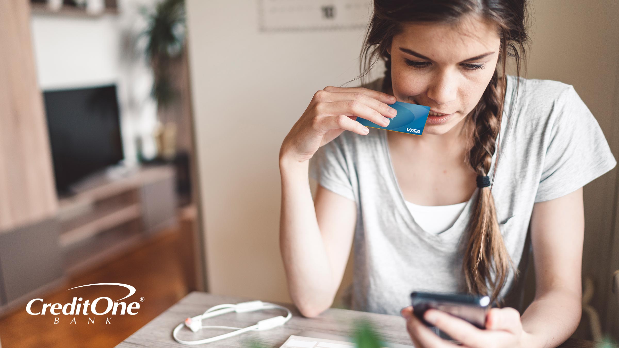 Seated young woman gently biting a credit card while looking at her smart phone