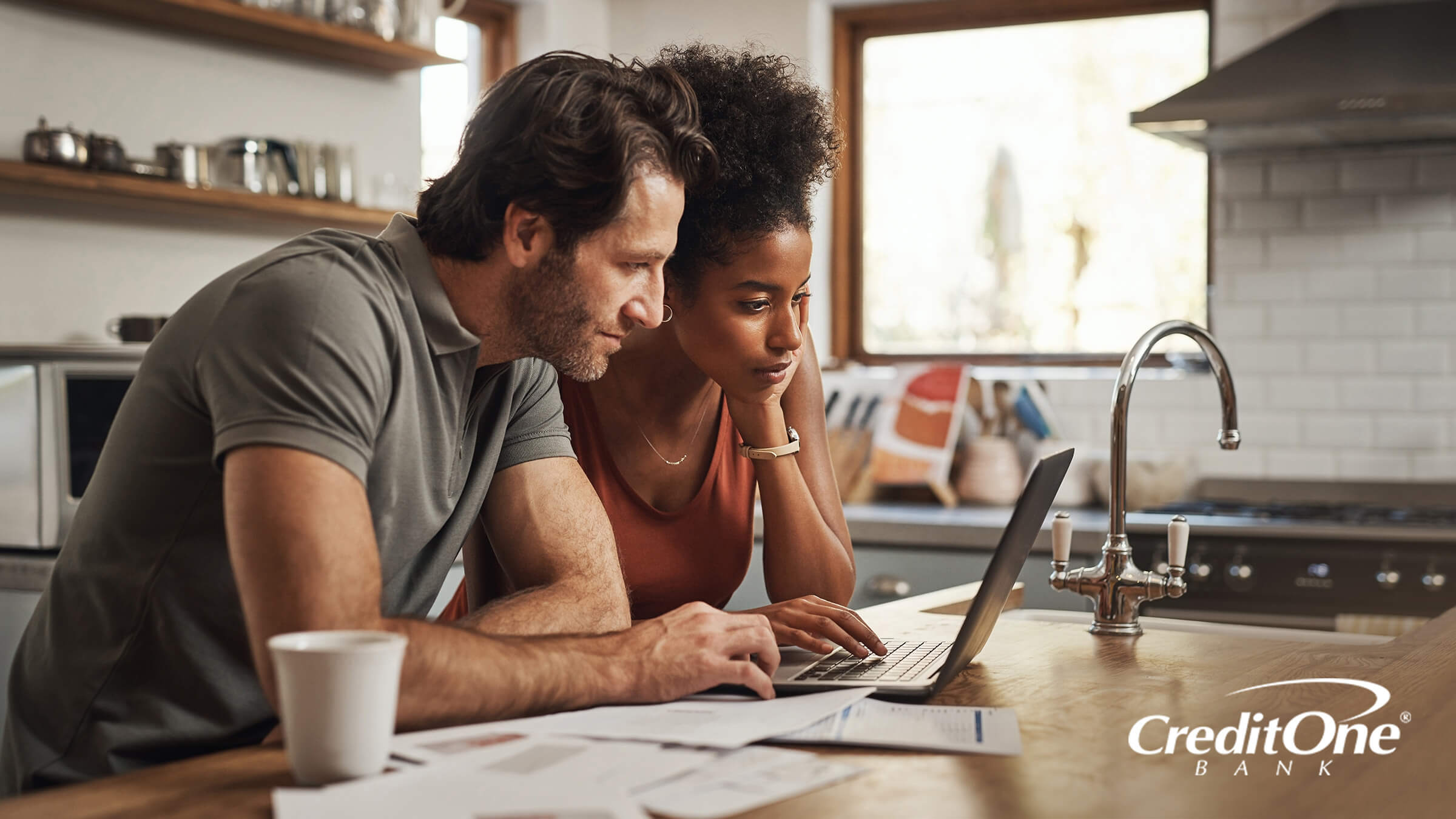 A man and woman browse on their laptop with financial paperwork on the countertop next to them, reviewing their finances for ways to protect their credit scores during a recession.