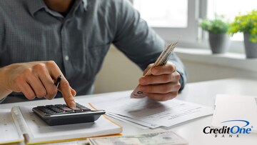 A man’s hands are shown holding a stack of cash while using a calculator, possibly figuring out whether to pay his credit card bill early.