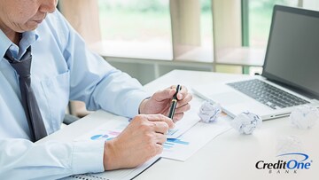 A businessperson looks worried as they sit at their desk and look over financial documents for their business — several of them are crumpled up into balls.