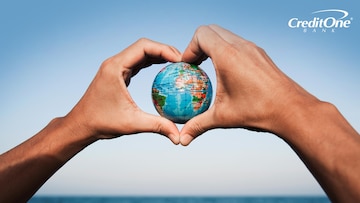 A man’s heart-shaped hands hold a globe in front of the ocean, representing a sustainable vacation