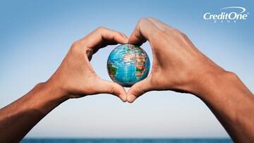 A man’s heart-shaped hands hold a globe in front of the ocean, representing a sustainable vacation