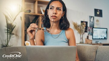 A woman sits on a couch with her laptop and ponders different factors before opening a certificate of deposit.