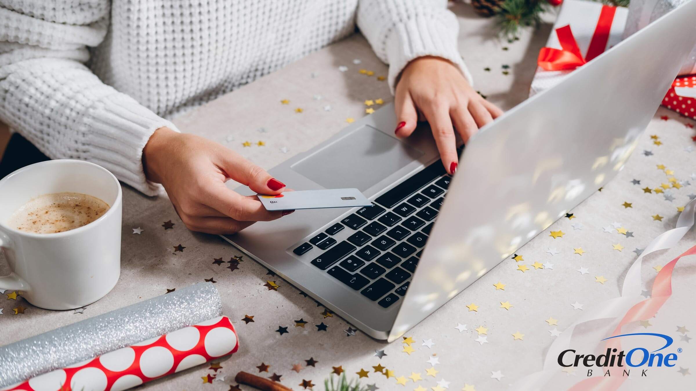 A woman’s hands are seen using her laptop and holding a credit card as she does her holiday shopping online with wrapping paper on the desk.