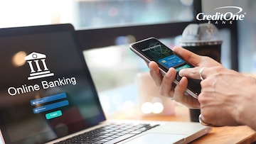 A man’s laptop screen shows his online banking website while his hands are seen using his banking mobile app at a coffee shop.