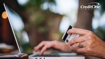 A close-up shot of a woman holding her credit card while also typing on her laptop, logging in to her online account to see what her outstanding balance is.