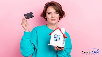 A young woman holds her credit card in one hand and a wooden model house in the other, representing the concept of paying a mortgage with a credit card.