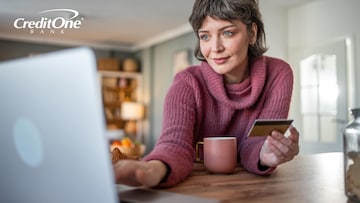 A smiling woman with a plum-colored sweater and matching coffee cup holds her credit card in one hand while using her laptop, possibly managing her credit card account online.
