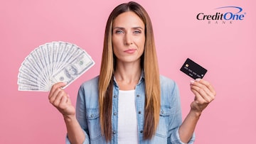 A woman holds up several $100 bills in her right hand and a credit card in her left, with a indecisive expression. Perhaps she is currently deciding whether cash or card is better.