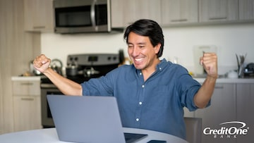 A man sitting at the kitchen table with his laptop smiles broadly and puts both fists in the air as if he’s celebrating his improved credit score.