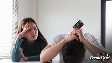 A couple sits at a table, the woman looking at the man with concern and frustration. The man has his face against the tabletop in shame, resting his hands on the back of his head while holding up a credit card. Perhaps he had been hiding his debt from loved ones.
