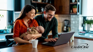 A couple reviews their finances on a laptop at the kitchen table, with the mother holding their newborn. They may be working on the family’s financial planning.