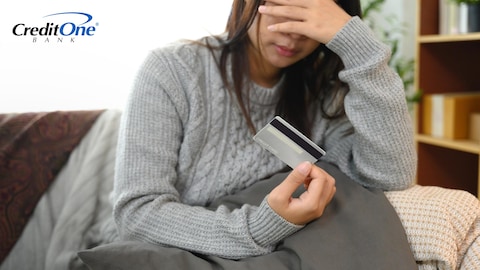 A young woman covers her face while sitting on her couch and holding a credit card in one hand, indicating that she may be confused or stressed about her credit.