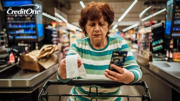 A senior woman checks her phone and receipt as she pushes her grocery cart past the store’s checkout counter with a concerned look. She may be worried about her current debt and the rising cost of groceries.