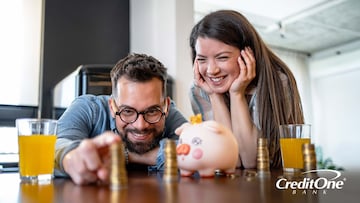 A happy couple is playing with stacks of coins on the table beside a piggy bank, separating their savings into categories like you would with both a high-yield and a traditional savings account.