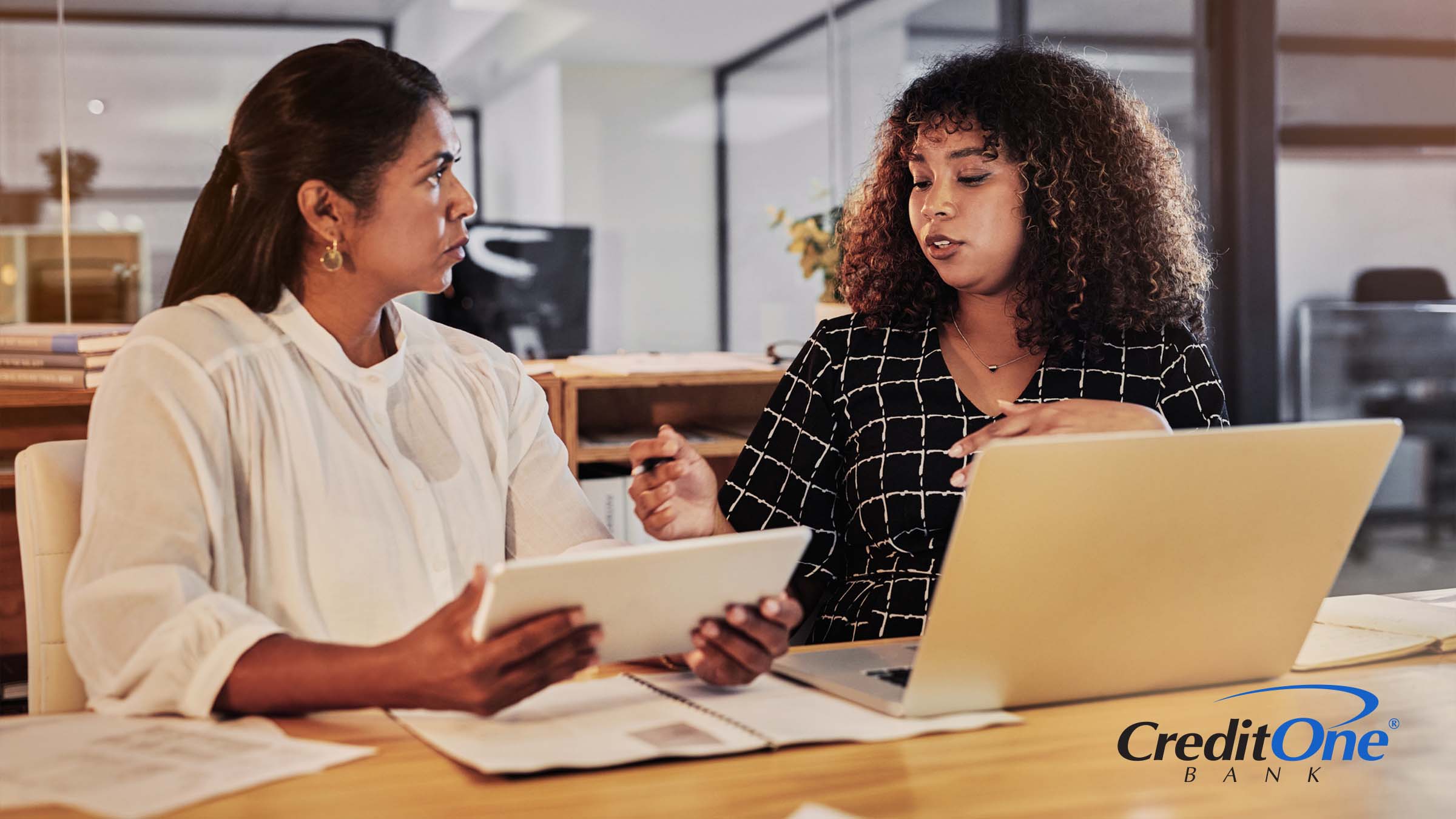 Two women have a discussion while sitting at a table. One has a laptop open while speaking and the other is listening carefully while holding on to her tablet. They may be discussing the differences between traditional and AI-based financial advice.