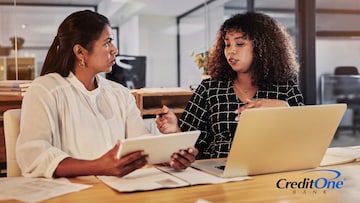 Two women have a discussion while sitting at a table. One has a laptop open while speaking and the other is listening carefully while holding on to her tablet. They may be discussing the differences between traditional and AI-based financial advice.