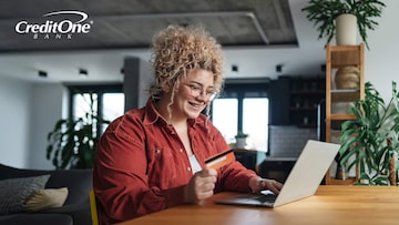 A smiling young woman uses her laptop at the table while holding her credit card in one hand, which may be a secured credit card to help her build credit.