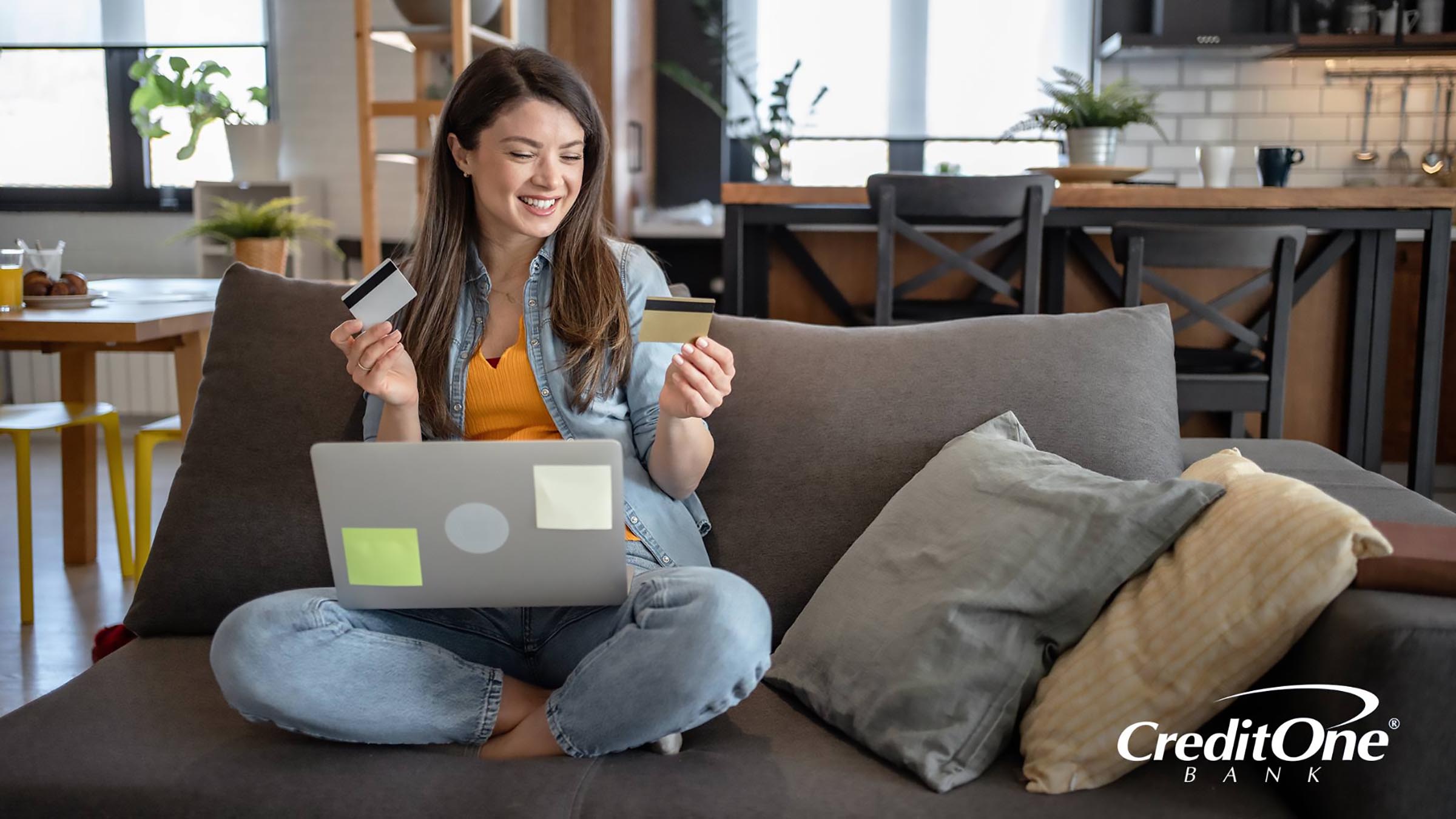 A young woman sits on her couch while happily comparing two credit cards, one in each hand. One may be a secured credit card and the other is her newly earned unsecured card.