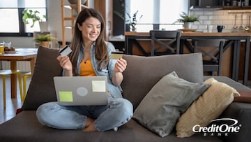 A young woman sits on her couch while happily comparing two credit cards, one in each hand. One may be a secured credit card and the other is her newly earned unsecured card.