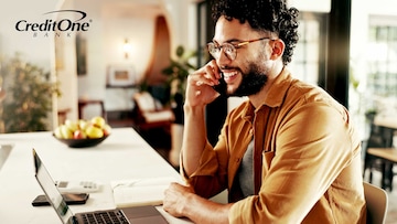 A young man sitting at a counter talks on his phone while smiling as if he’s getting good news, like an update to his credit score or loan application.