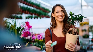 A smiling woman holds a potted plant in a retailer’s garden section, while presenting her store credit card for payment. 