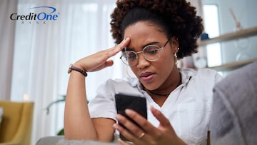 A woman rests on her sofa while looking into her phone with a frustrated look. She may be annoyed after receiving an AI-generated scam text.