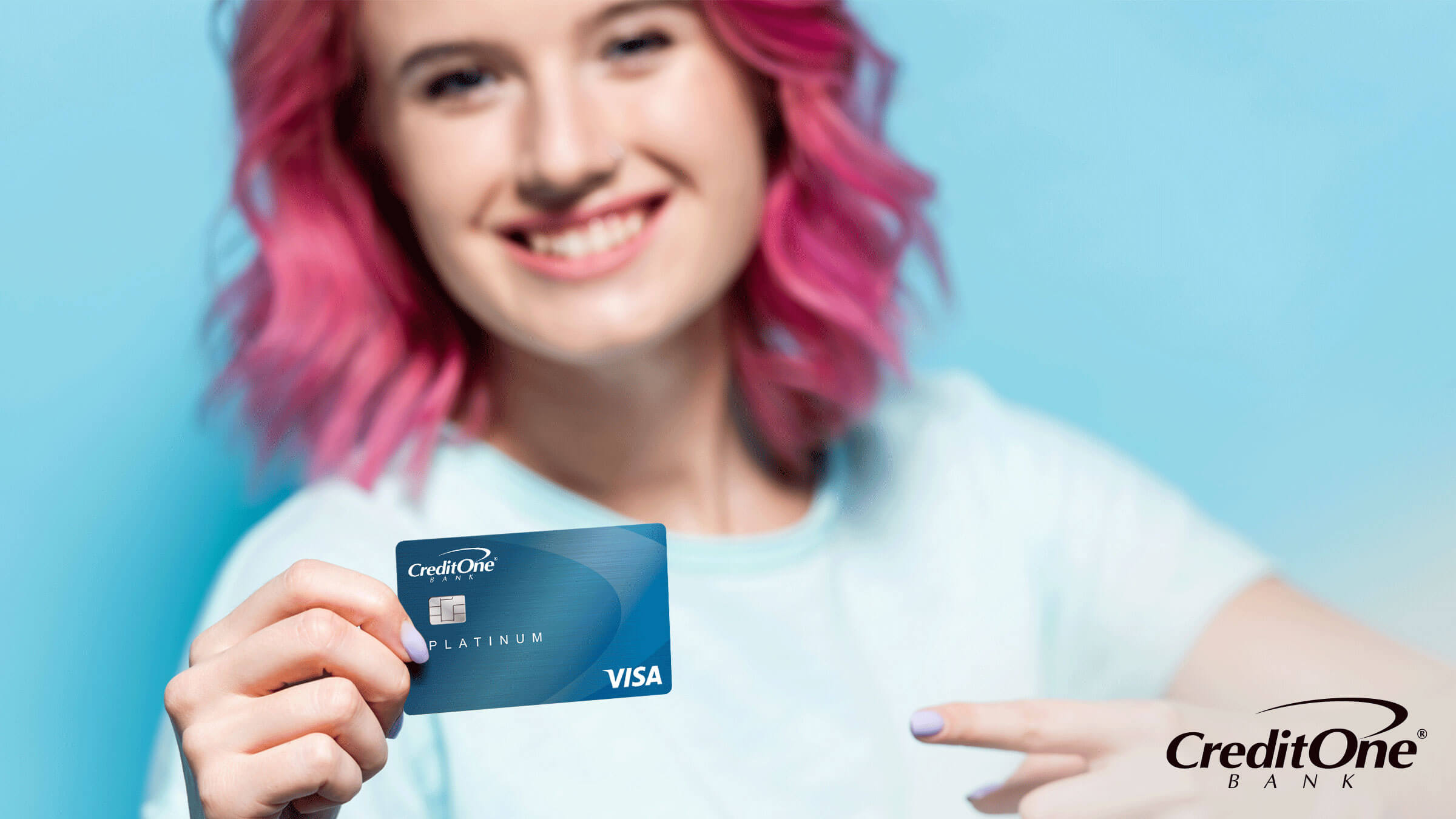 A young person with pink hair smiles as they hold their credit card up with one hand while pointing to it with the other.
