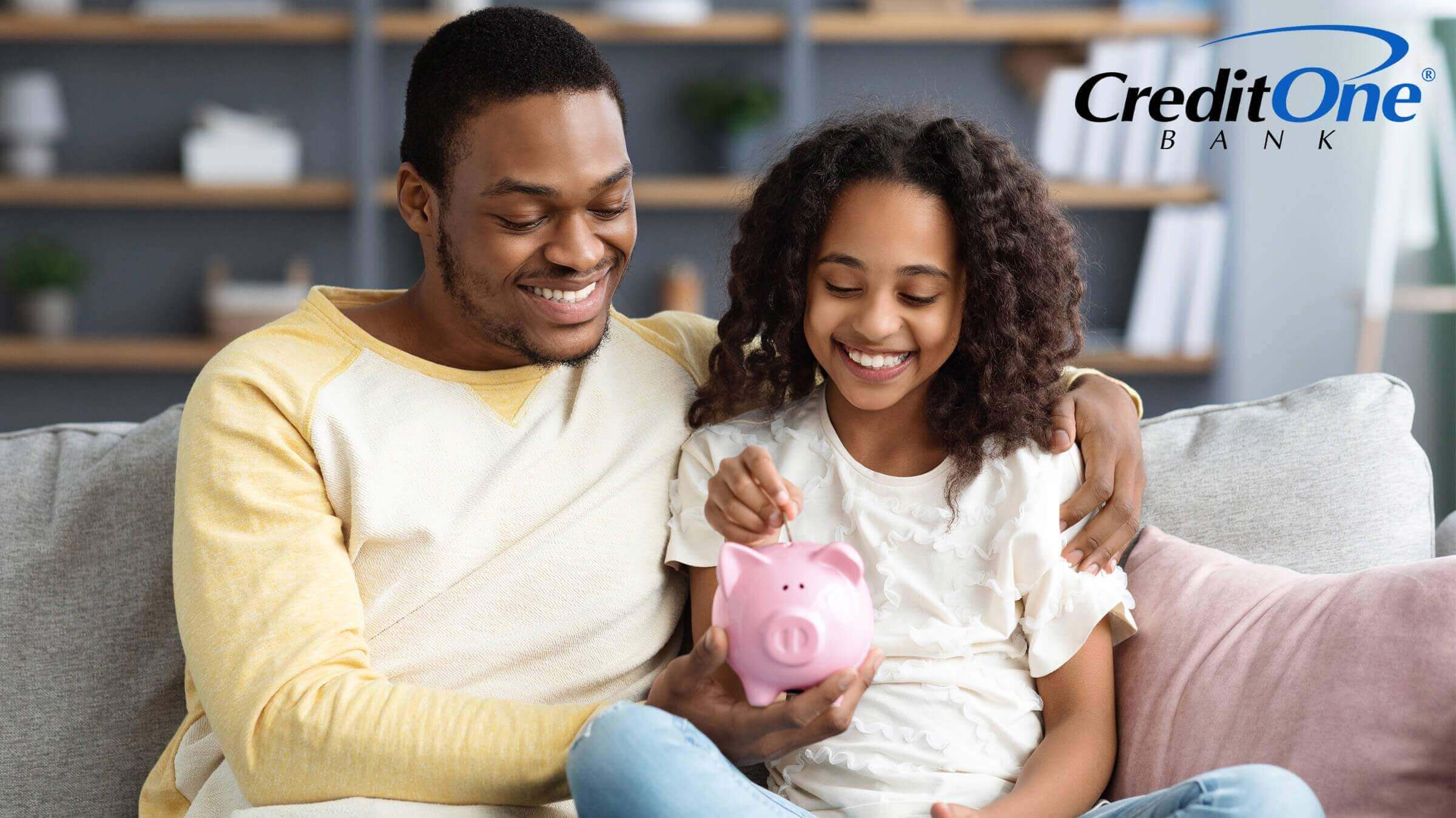 A father teaches his daughter about financial literacy as they sit on the couch and put coins in her piggy bank