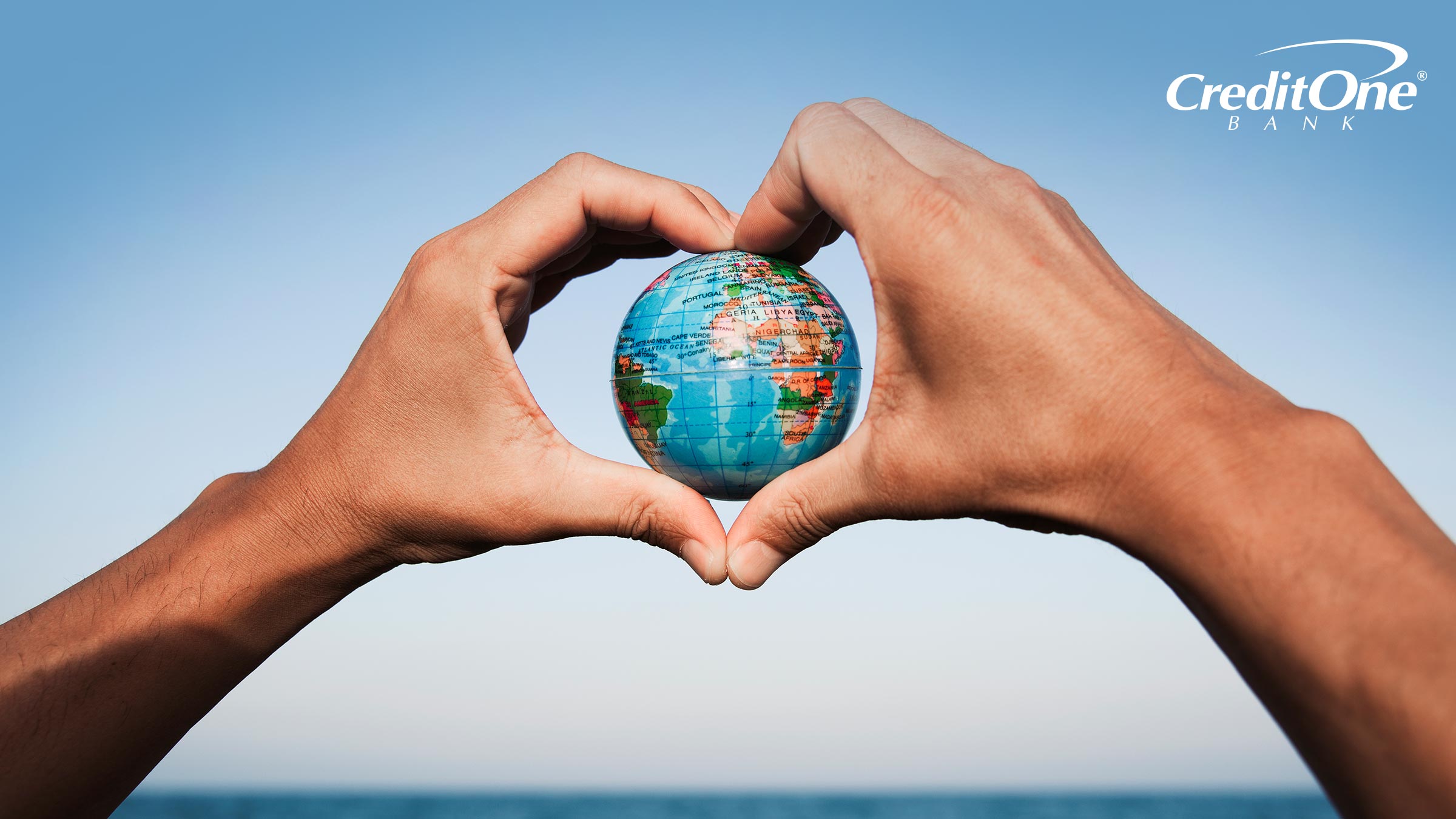 A man’s heart-shaped hands hold a globe in front of the ocean, representing a sustainable vacation A man’s heart-shaped hands hold a globe in front of the ocean, representing a sustainable vacation
