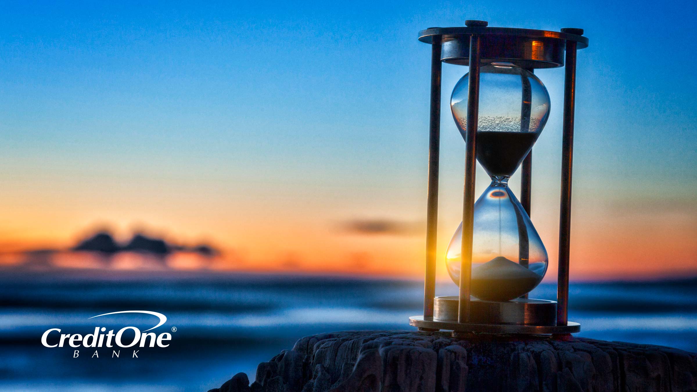 An hour glass rests on a rock with the sun setting in the background, symbolizing the passage of time and a credit card’s expiration date.
